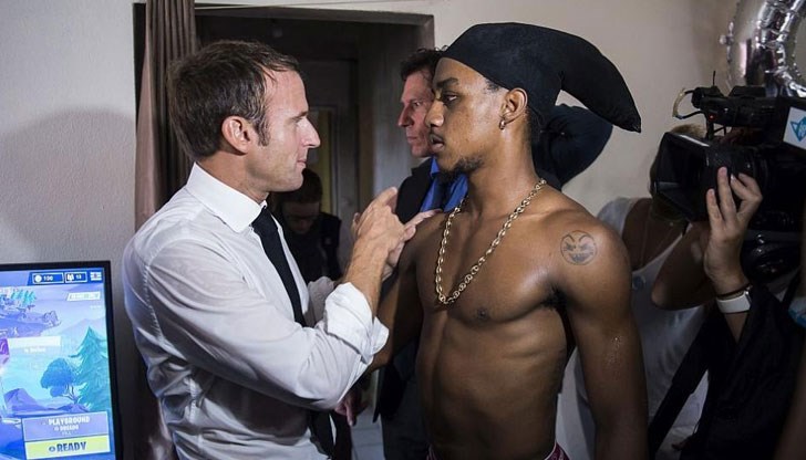 Френският президент позира усмихнат между двама младежи, единият от които прави вулгарен жест