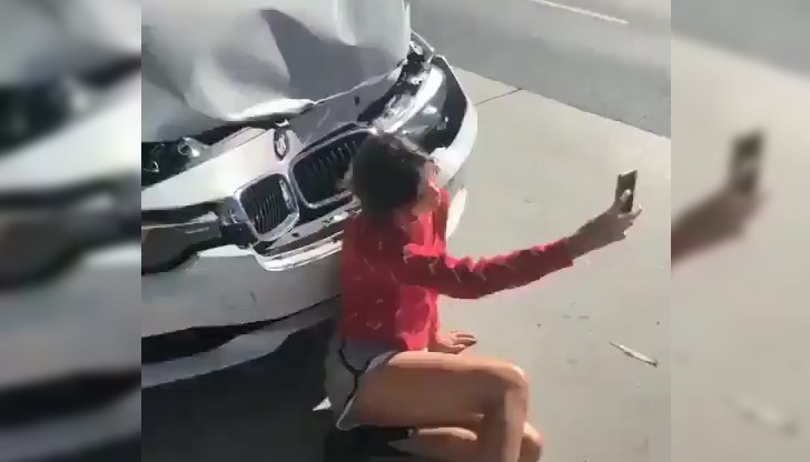 Тя е седнала на земята пред размазаната предница на автомобила и си прави снимка за спомен от инцидента